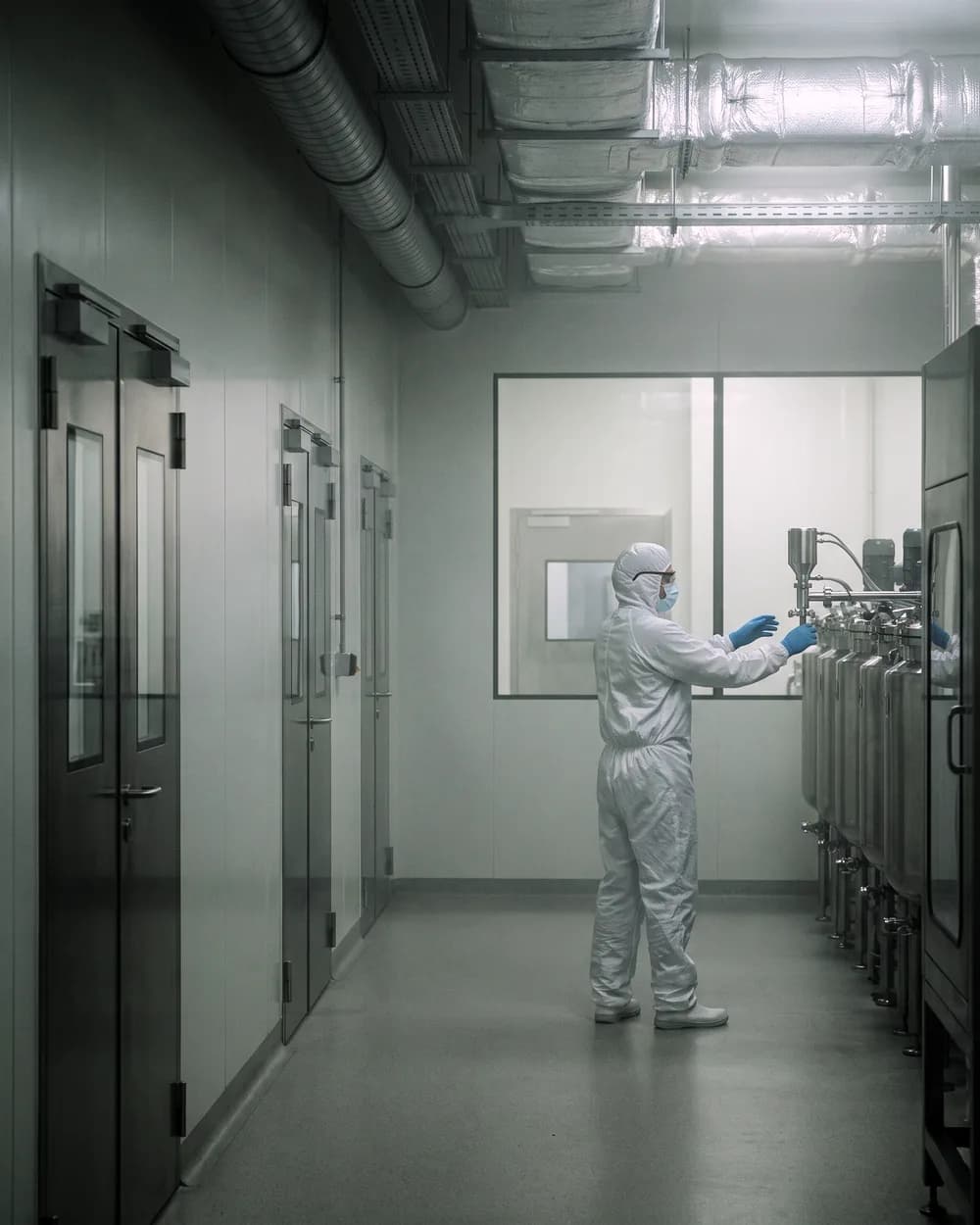 Illustrative frame: a pharmaceutical clean room corridor, sealed doors along one side and an operator in full coverall working a filling line at the far end.