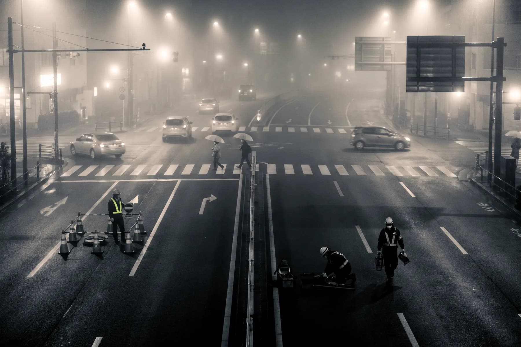 Illustrative frame: an urban road junction seen from an elevated mast at night in rain, lanes of wet asphalt under signal gantries with a few vehicles crossing.